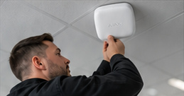 Engineer inspecting a white Ajax EN54 ceiling detector, from the approved artwork